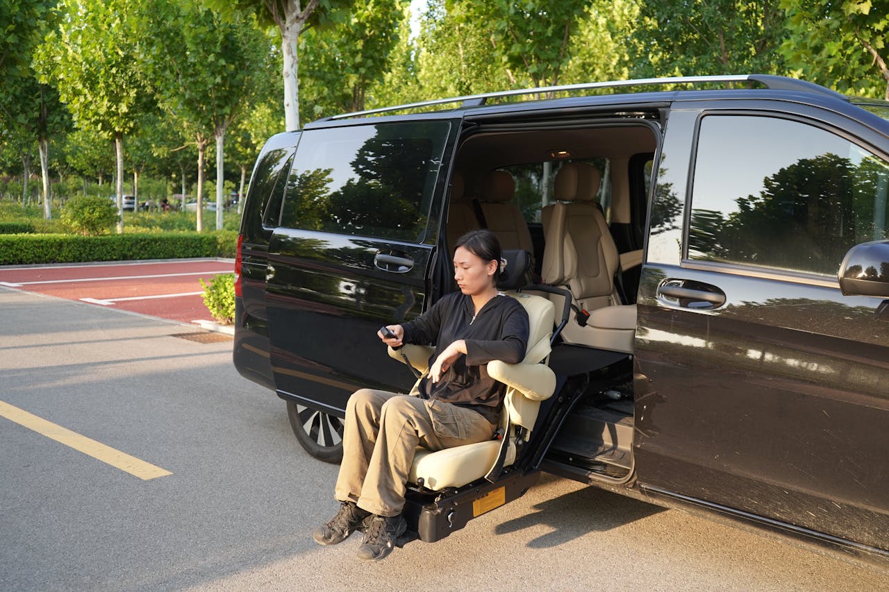 Woman using a seat lift in a black van at a parking lot with trees in the background.